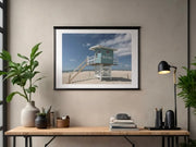 Framed French beach with lifeguard tower scene print on a wall above a desk with plants and a lamp.