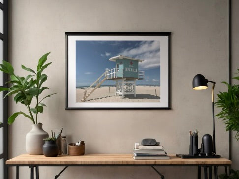 Framed French beach with lifeguard tower scene print on a wall above a desk with plants and a lamp.
