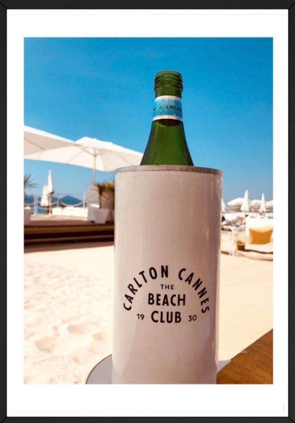 Bottle of sparkling water in a cooler with 'Carlton Cannes The Beach Club' branding on a beach setting.