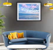 Modern living room with a blue sofa, yellow pillows, a plant, and framed blue Paradise now open artwork on a gray wall.