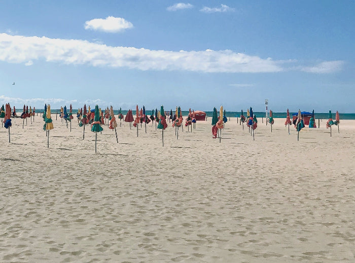 A wide sandy beach in Deauville, France with its signature colourful parasols arranged in rows under a blue sky with white clouds, with the ocean visible in the distance.