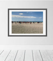 A framed beach scene print hanging on a white wall above white wooden floorboards. The artwork shows a sandy beach in Deauville, France with colourful parasols under a blue sky with clouds.