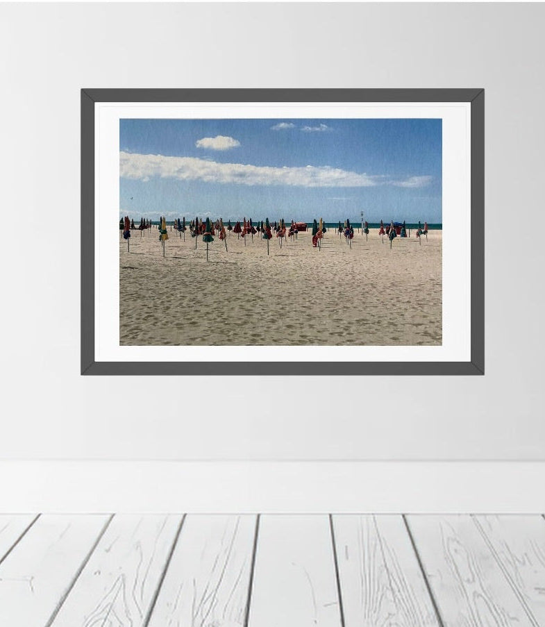 A framed beach scene print hanging on a white wall above white wooden floorboards. The artwork shows a sandy beach in Deauville, France with colourful parasols under a blue sky with clouds.