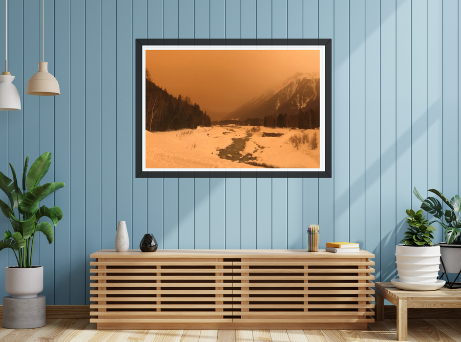 A framed landscape art print of a snowy mountain valley under a warm orange sky, displayed on a light blue wood-paneled wall above a wooden console table in a styled room.