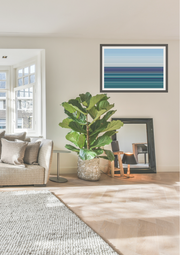 A framed abstract art print with horizontal stripes in shades of blue, teal, and gray, displayed on a beige wall in a modern living room setting next to a potted fiddle-leaf fig plant.