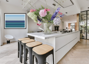 A modern kitchen island with a white marble countertop, three black bar stools with light wood seats, and a large glass vase filled with colorful flowers on top. The kitchen has light wood herringbone flooring, white walls, and a framed blue ocean print on the left wall.