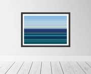 A framed horizontal art print featuring blue and teal ocean waves, displayed on a light gray wall above a white wood plank floor.