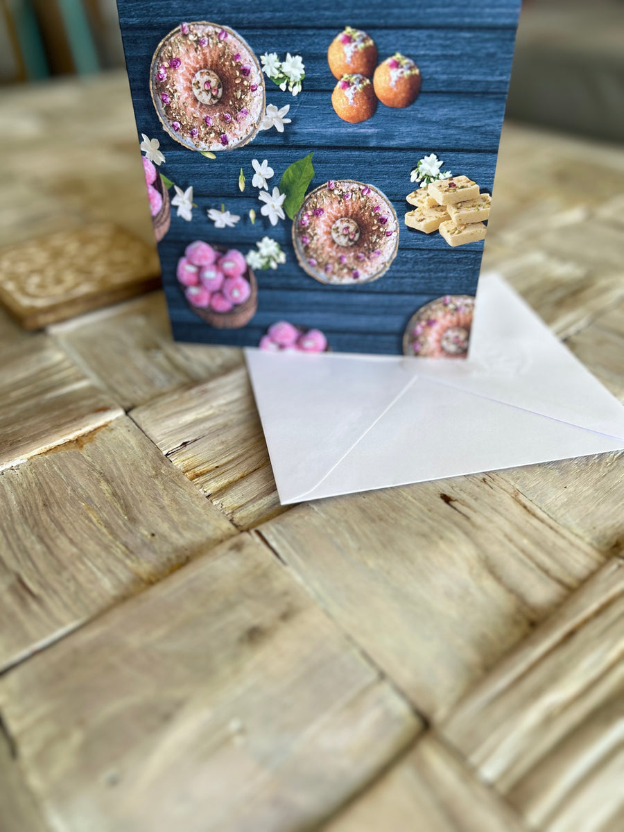 A vibrant greeting card featuring traditional Indian sweets like ladoos, barfi, and jalebis on a rich teal wooden background. Decorated with jasmine flowers and intricate details, this seamless pattern adds a festive, cultural touch, perfect for celebrations.