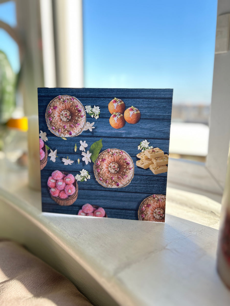 A vibrant greeting card featuring traditional Indian sweets like ladoos, barfi, and jalebis on a rich teal wooden background. Decorated with jasmine flowers and intricate details, this seamless pattern adds a festive, cultural touch, perfect for celebrations.
