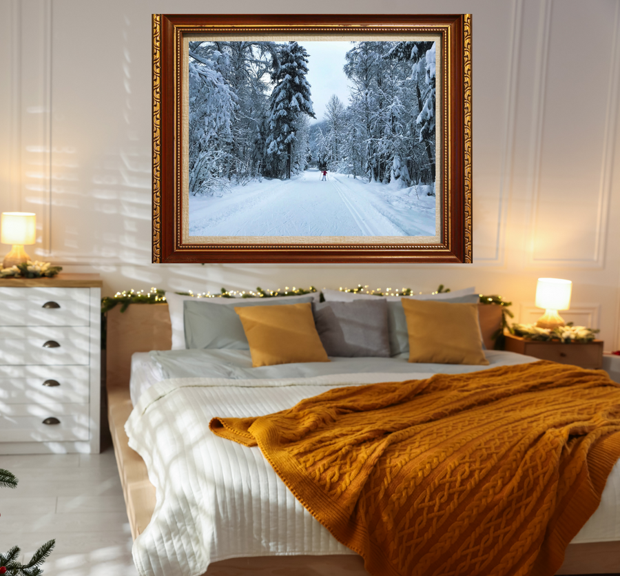 A framed winter landscape painting hangs on a white paneled wall above a neatly made bed in a warmly lit bedroom. The artwork shows a snow-covered path through a forest, and the room includes bedside tables, lamps, pillows, and a mustard blanket.