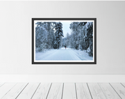A framed winter landscape photograph hanging on a light gray wall above white wooden floorboards. The print shows a snow-covered forest path with snow-covered trees on both sides.