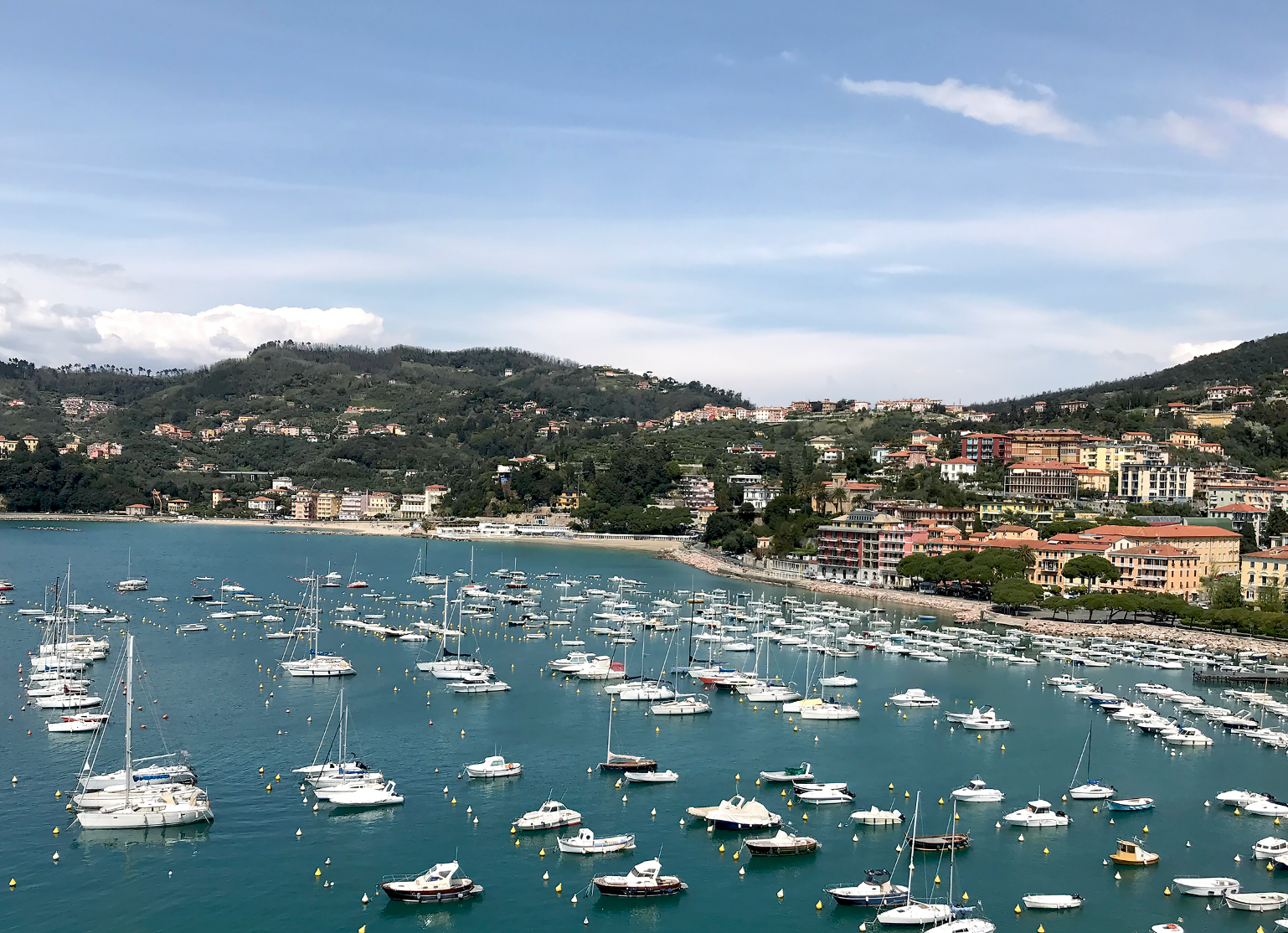 A scenic coastal harbor on the Italian Riviera filled with numerous white sailboats and yachts on turquoise-blue water, with a hillside town and buildings rising behind the marina under a partly cloudy blue sky.