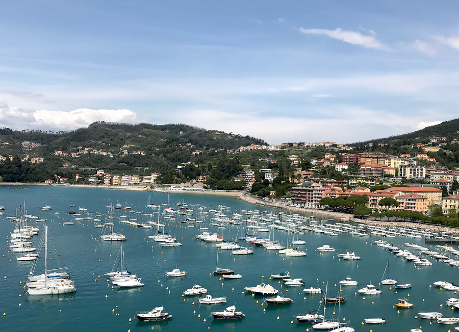 A scenic coastal harbor on the Italian Riviera filled with numerous white sailboats and yachts on turquoise-blue water, with a hillside town and buildings rising behind the marina under a partly cloudy blue sky.