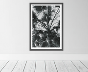 A framed black and white botanical art print of palm leaves, displayed on a light gray wall above white wooden floorboards.