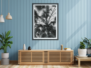 A framed black and white palm tree art print hangs on a light blue wood-paneled wall in a modern room setting with a wooden console table, potted plants, and pendant lights.