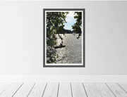 A framed landscape fine art print depicting a shimmering River Thames and a small sailboat, hung on a light gray wall above white wooden floorboards.
