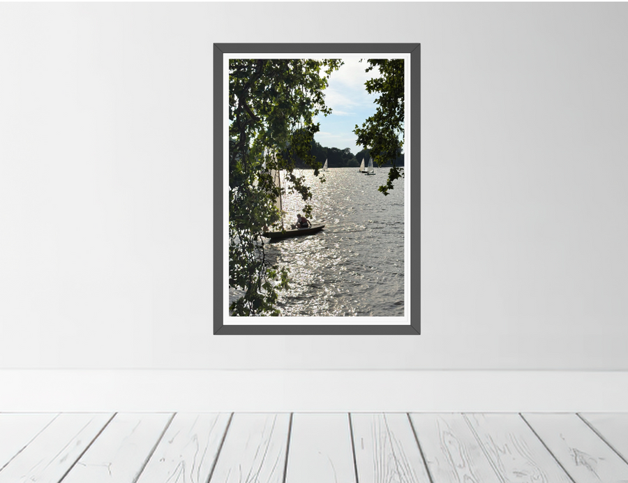 A framed landscape fine art print depicting a shimmering River Thames and a small sailboat, hung on a light gray wall above white wooden floorboards.
