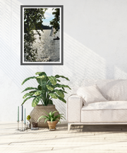 A framed vertical fine art photography print by Seafoam Atelier photographer, Samira Malik depicting a sailboat entering the frame of the river Thames with trees and shimmering water, styled in a bright, modern living room above a potted green plant and next to a light-colored sofa.