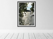 A framed vertical art print depicting the River Thames with a sailboat and tree branches in the foreground and shimmering water in the afternoon sun. Limited edition fine art print by Seafoam Atelier photographer, Samira Malik