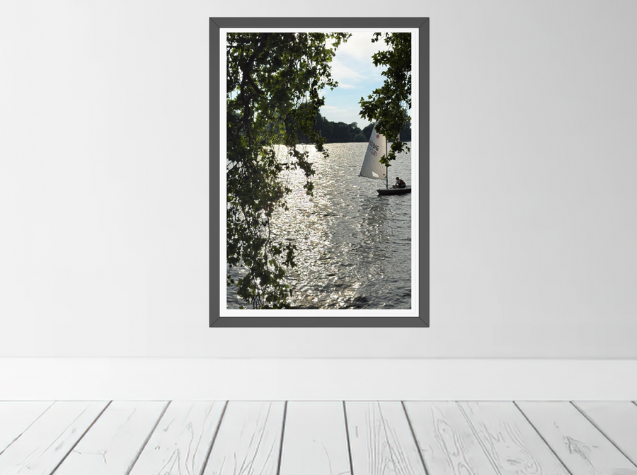 A framed vertical art print depicting the River Thames with a sailboat and tree branches in the foreground and shimmering water in the afternoon sun. Limited edition fine art print by Seafoam Atelier photographer, Samira Malik