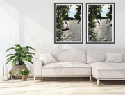 A modern living room scene with a light beige sectional sofa, a potted green plant, and two framed vertical landscape prints from the River Thames Shimmer series, hanging on the wall above the sofa. The prints depict a reflective river Thames scene with trees and dark shoreline and are photographed by Seafoam Atelier photographer, Samira Malik.  