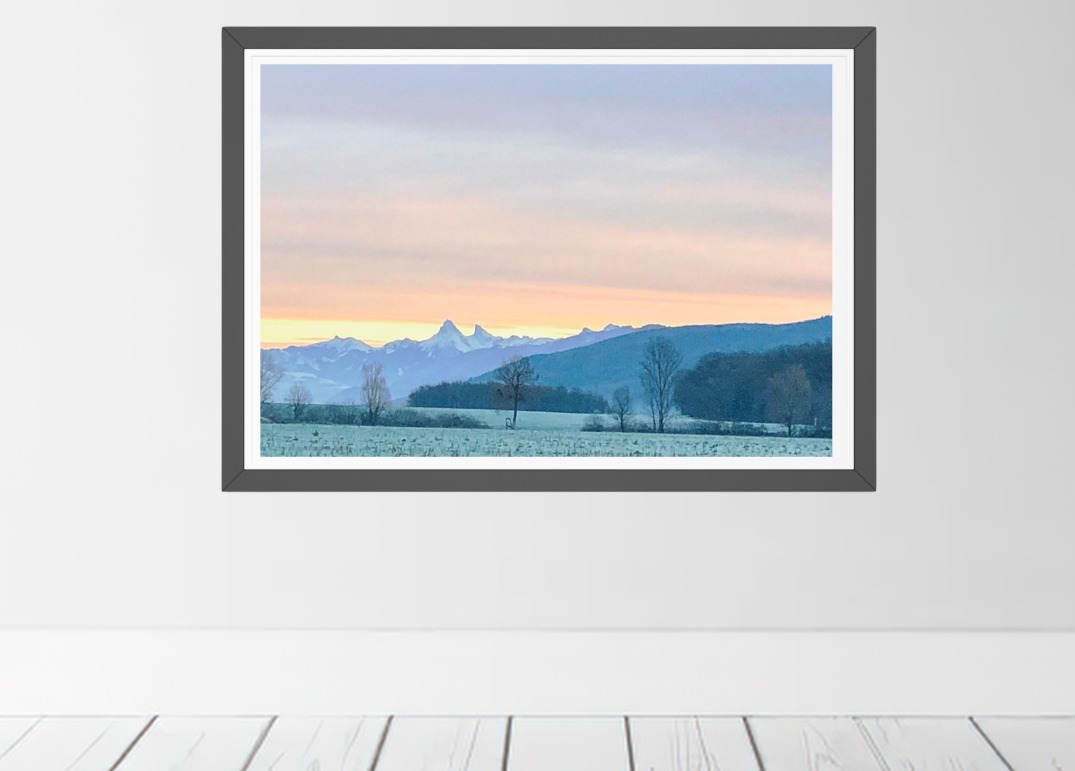 A framed landscape art print featuring a scenic mountain valley at sunset, displayed on a light gray wall above white wooden floorboards.