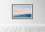 A framed landscape art print featuring a scenic mountain valley at sunset, displayed on a light gray wall above white wooden floorboards.