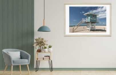 Framed photograph of a French beach lifeguard hut scene on a wall above a chair and side table.