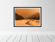 A framed landscape art print showing a snowy mountain valley in Chamonix with the air full of orange sand from the Sahara desert. Displayed on a light gray wall above white wooden floorboards.