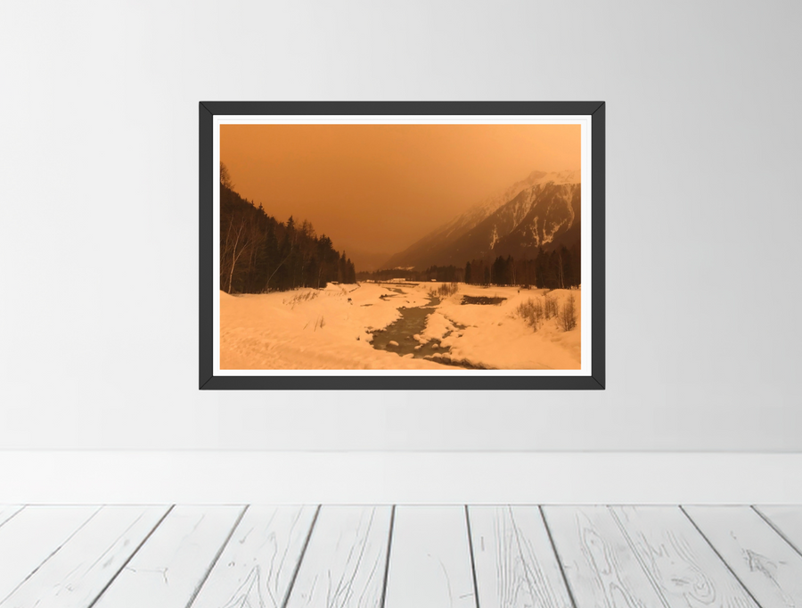 A framed landscape art print showing a snowy mountain valley in Chamonix with the air full of orange sand from the Sahara desert. Displayed on a light gray wall above white wooden floorboards.