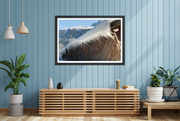 A framed landscape art print of snowy mountains and a wooden structure, displayed on a blue paneled wall in a modern living room setting.