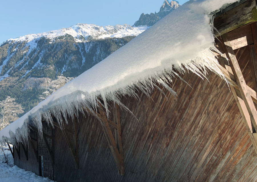 A rustic wooden building with a snow-covered roof and long icicles hanging over the eaves, set against a mountain landscape under a clear blue sky.