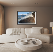A framed landscape art print of a icy chalet and snowy mountain peak under a blue sky, displayed on a taupe wall in a modern living room setting above a white sectional sofa.