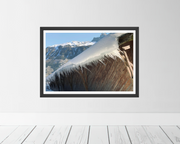 A framed art print depicting a snowy mountain landscape under a blue sky, mounted on a light gray wall above white wooden floorboards.
