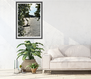 A framed vertical fine art photography print featuring a small sailboat on a shimmering River Thames. Photographed by Seafoam Atelier photographer, Samira Malik.  Styled on a white wall above a light-colored sofa and potted plants in a modern living room setting.
